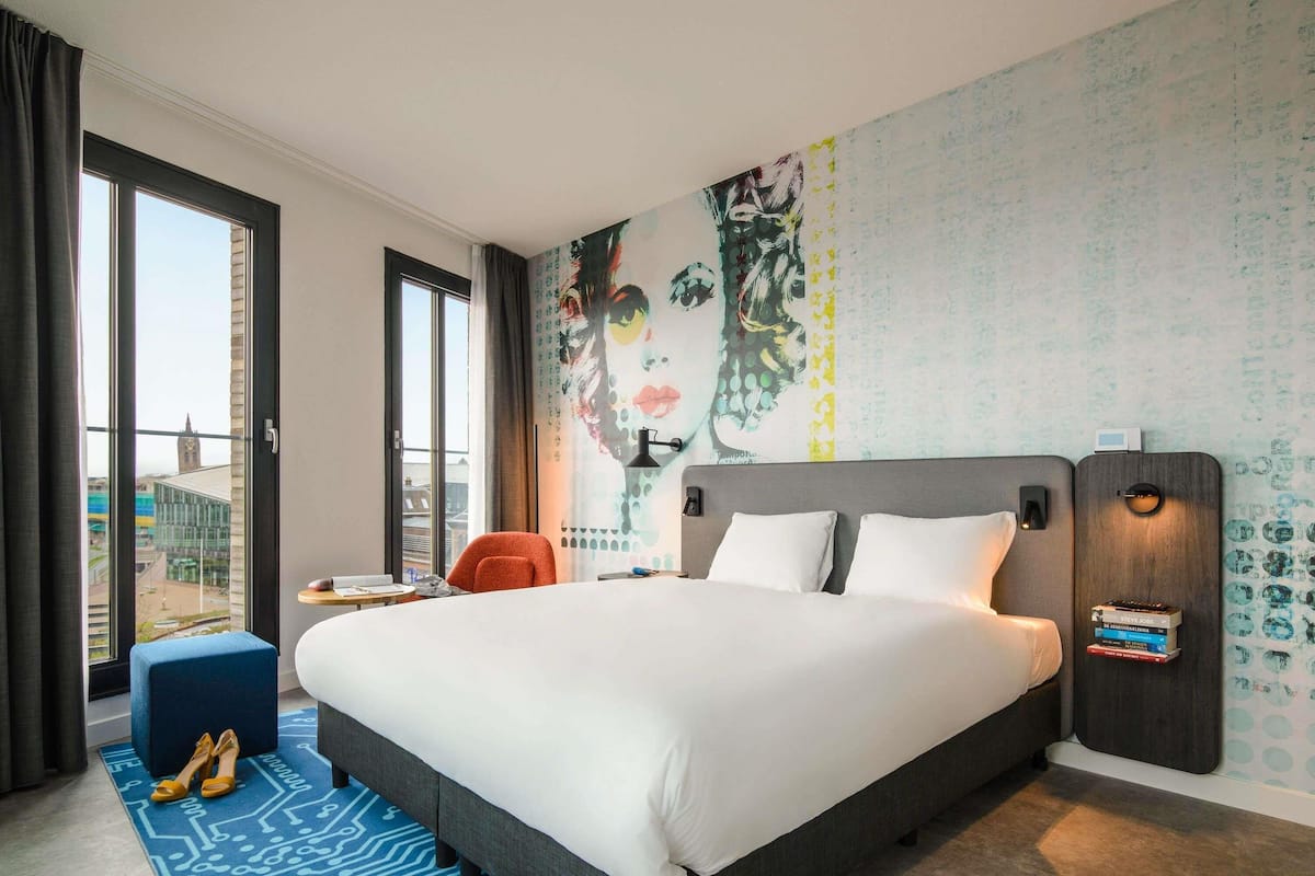 A hotel room with a large bed, a desk, a chair, a window with a view, and a wall with a mural.