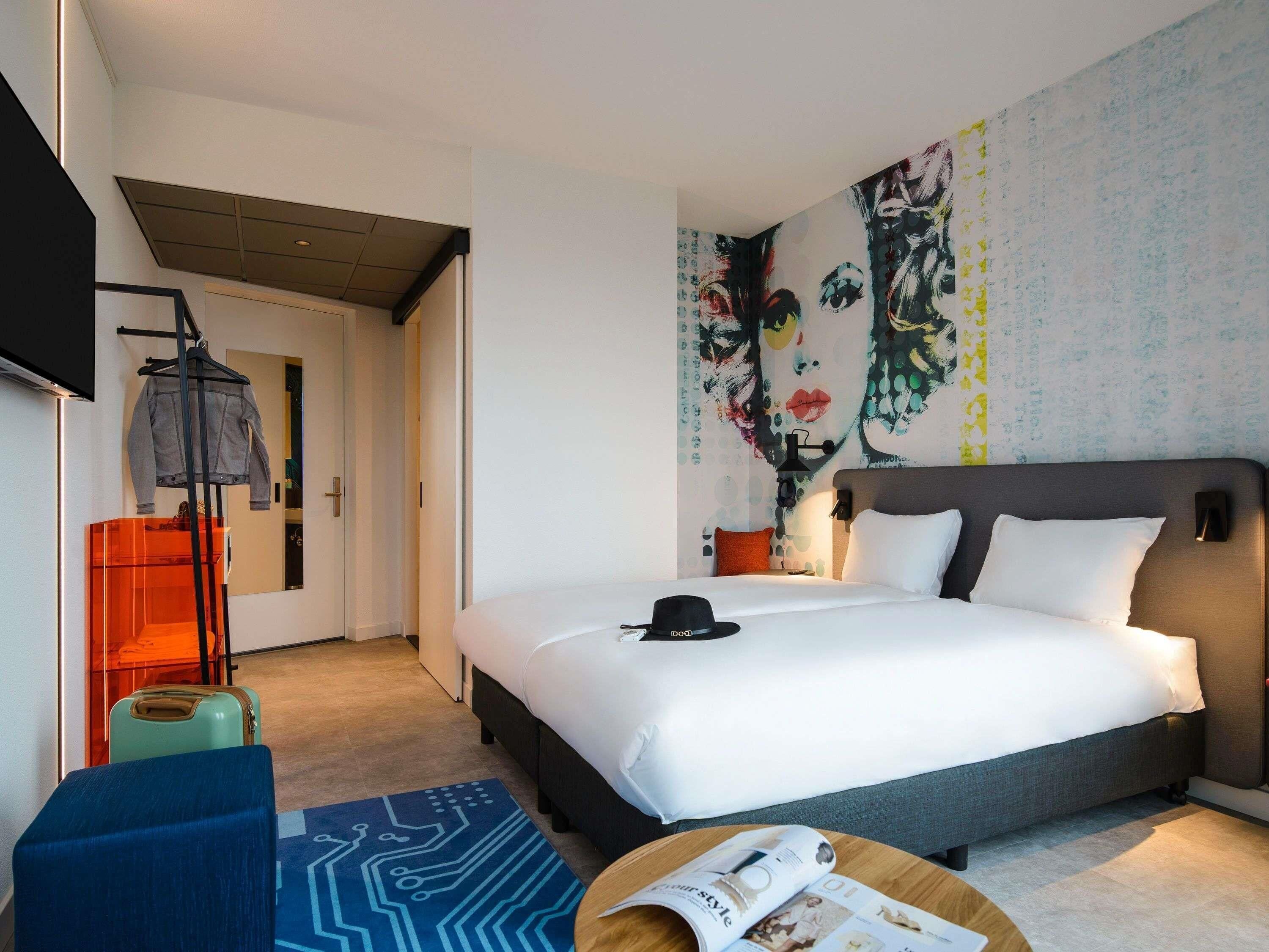 Une chambre d’hôtel moderne dotée d’un grand lit, d’un téléviseur à écran plat, d’un bureau avec une chaise et d’un mur orné d’un motif abstrait aux couleurs vives.