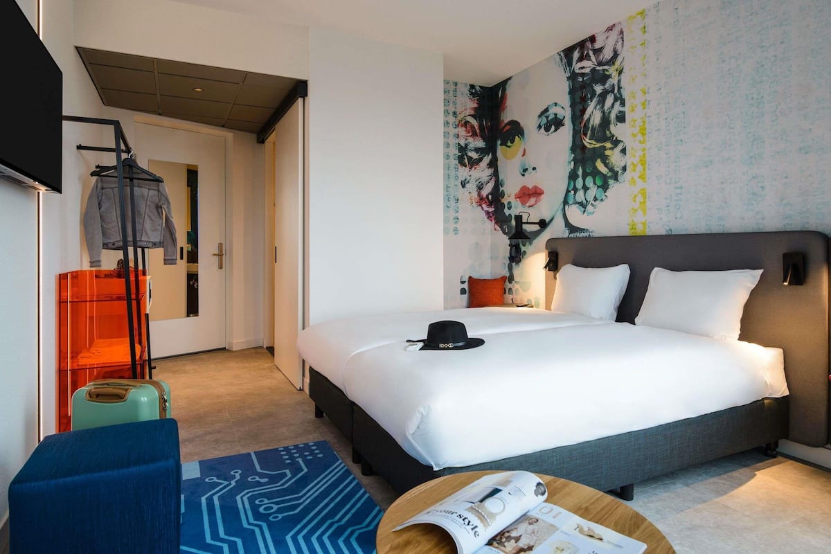Une chambre dâhĂŽtel moderne dotĂ©e dâun grand lit, dâun tĂ©lĂ©viseur Ă Ă©cran plat, dâun bureau avec une chaise et dâun mur ornĂ© dâun motif abstrait aux couleurs vives.