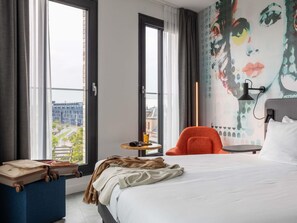 A hotel room with a bed, a chair, a small table with a lamp, and a large window with a view of buildings. - ibis Styles Delft City Centre (Delft)