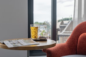A table with a glass of amber liquid, a bowl of food, and a magazine, set against a window with a view of buildings and a bridge. - ibis Styles Delft City Centre (Delft)