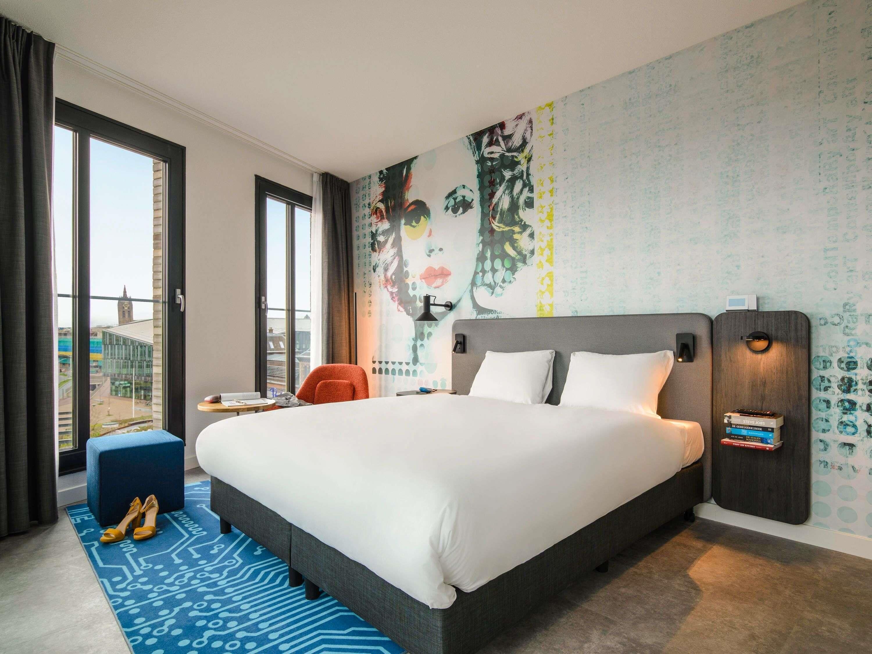 Une chambre d’hôtel avec un grand lit, un bureau, une chaise, une fenêtre donnant sur un beau paysage et un mur orné d’une fresque.