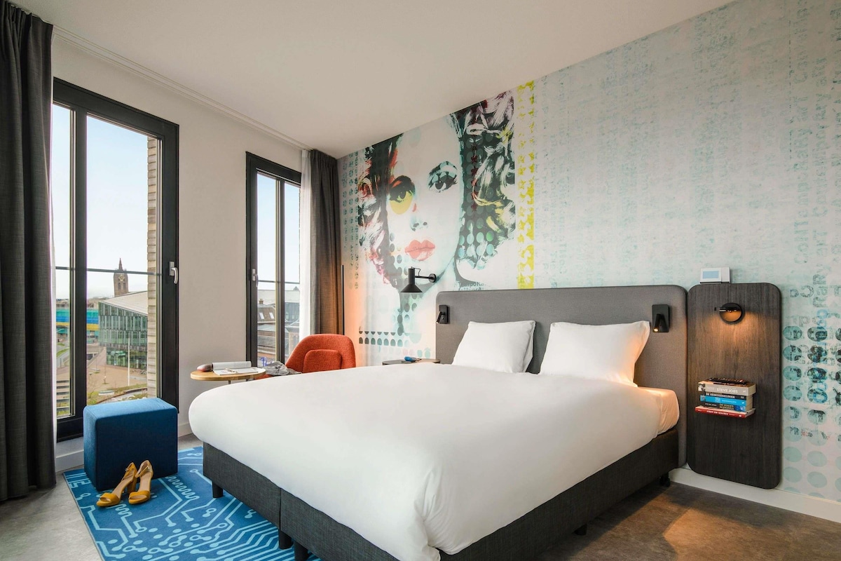 Une chambre d’hôtel avec un grand lit, un bureau, une chaise, une fenêtre donnant sur un beau paysage et un mur orné d’une fresque.