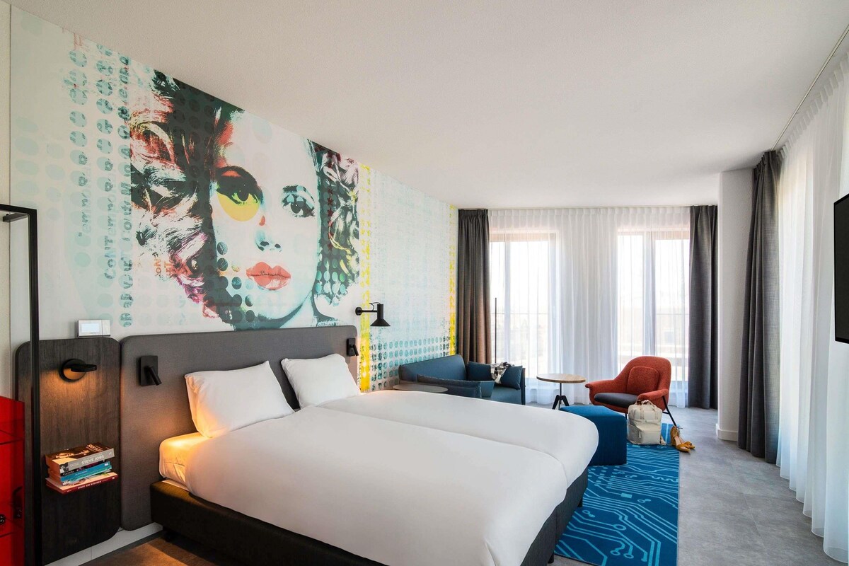 Une chambre d’hôtel dotée d’un grand lit, d’un coin salon avec un canapé et un fauteuil, d’une télévision et d’un grand tableau abstrait accroché au mur.