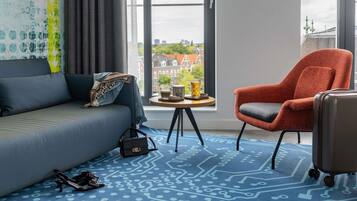 A modern hotel room with a sofa, armchair, and a view of a cityscape.