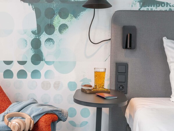 Une chambre d’hôtel avec un lit, une petite table sur laquelle se trouvent une boisson et un livre, un casque posé sur une chaise, et un mur orné d’un motif géométrique.