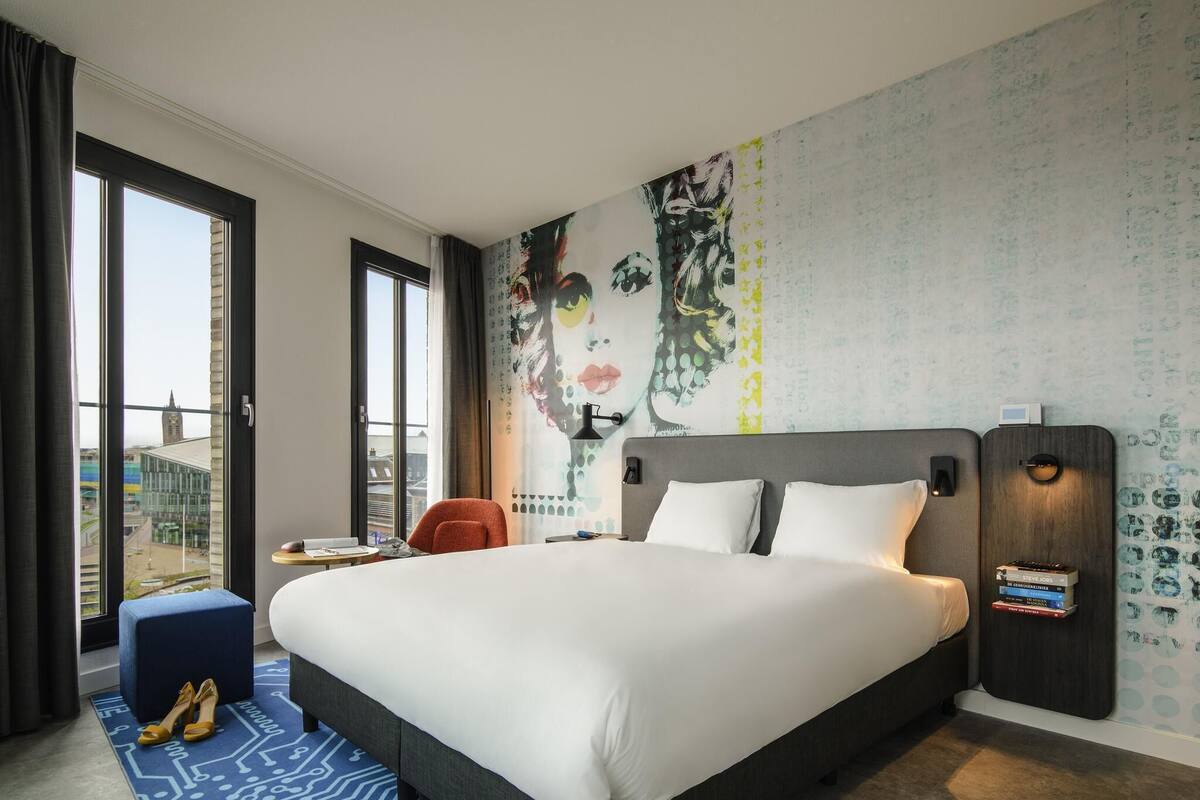 Ein Hotelzimmer mit einem großen Bett, einem Sessel, einem kleinen Tisch und Blick auf die Stadt durch große Fenster.