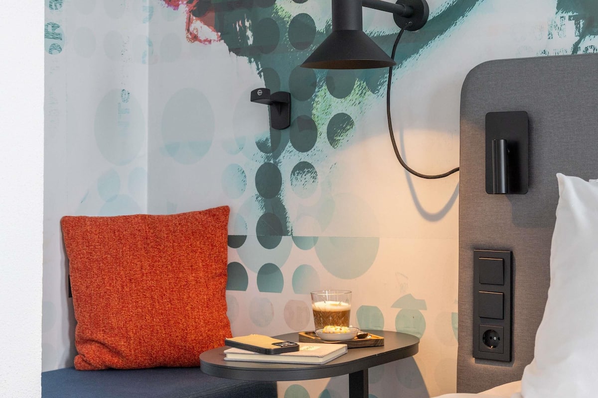 Une chambre dâhĂŽtel avec une table ronde, un coussin rouge, un verre de biĂšre, un carnet et un tĂ©lĂ©phone intelligent.