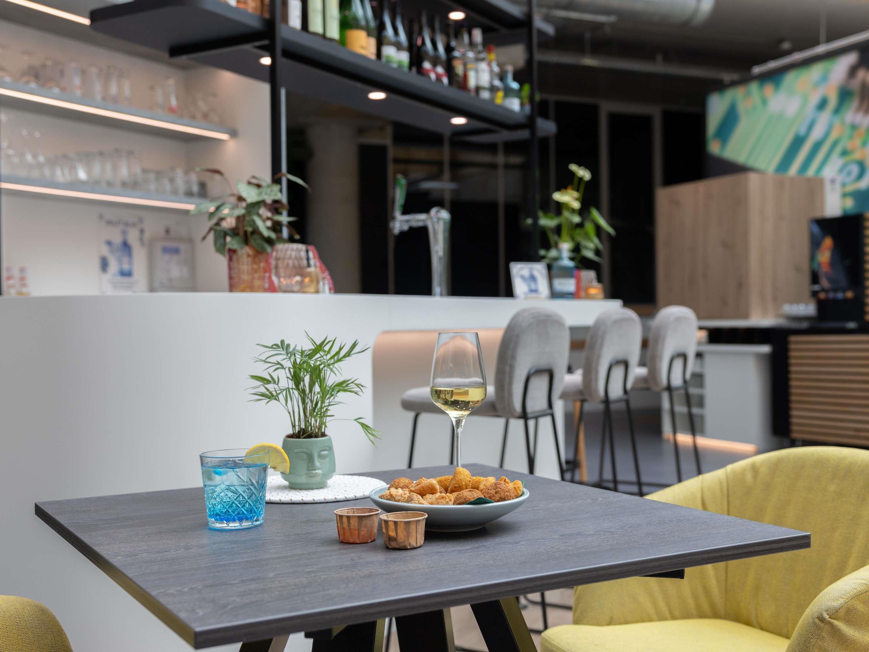 A modern bar area with a table set for two, featuring a glass of white wine, a blue drink, and a bowl of fried snacks.