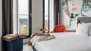 Une chambre d’hôtel avec un lit, une valise, une chaise, une petite table et un mur orné d’une fresque colorée.