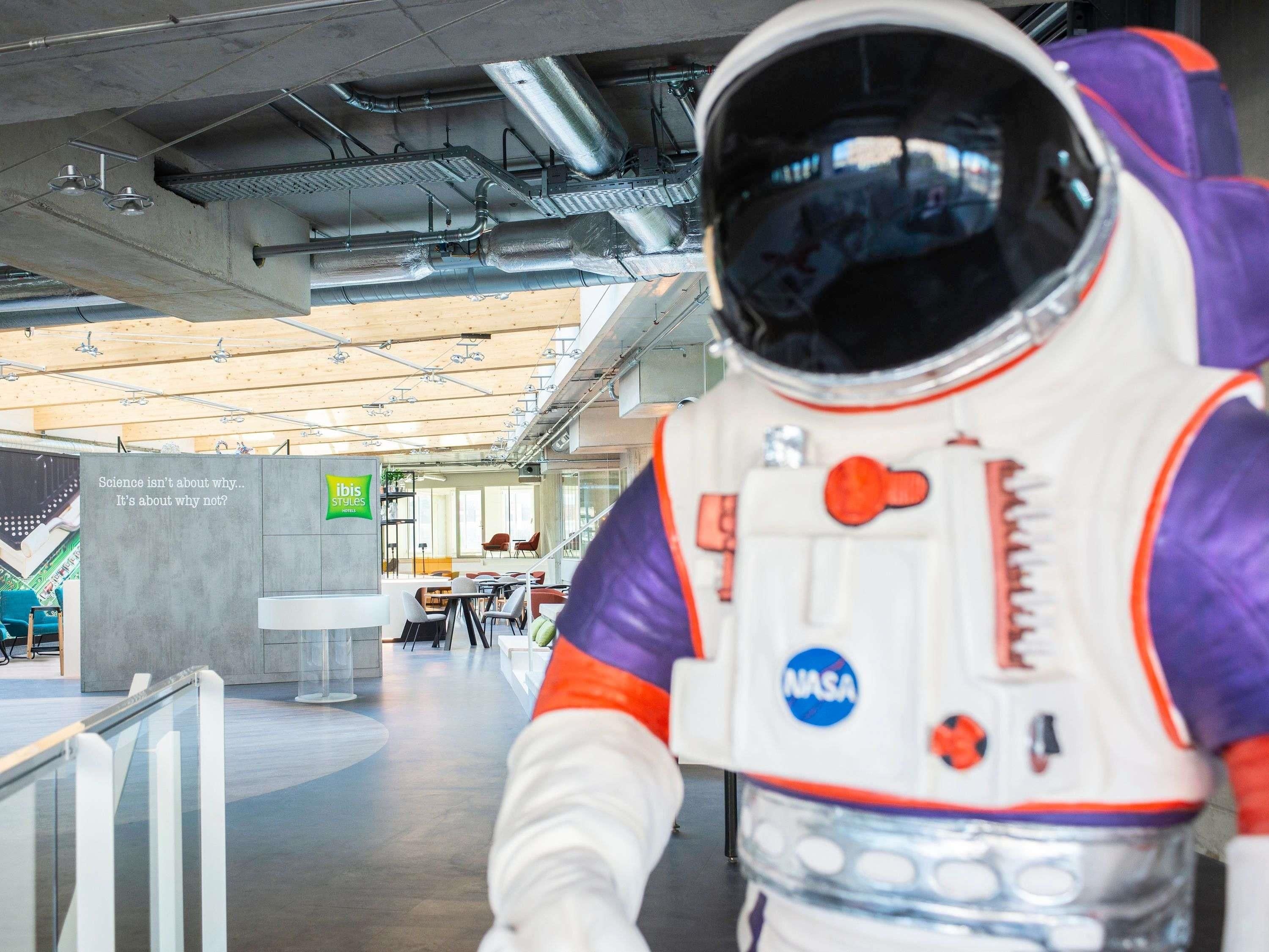 An astronaut figure in a modern indoor space with visible ceiling pipes and a sign for 'Ibis Styles Hotel'.