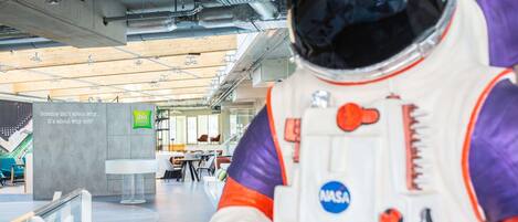 An astronaut figure in a modern indoor space with visible ceiling pipes and a sign for 'Ibis Styles Hotel'.