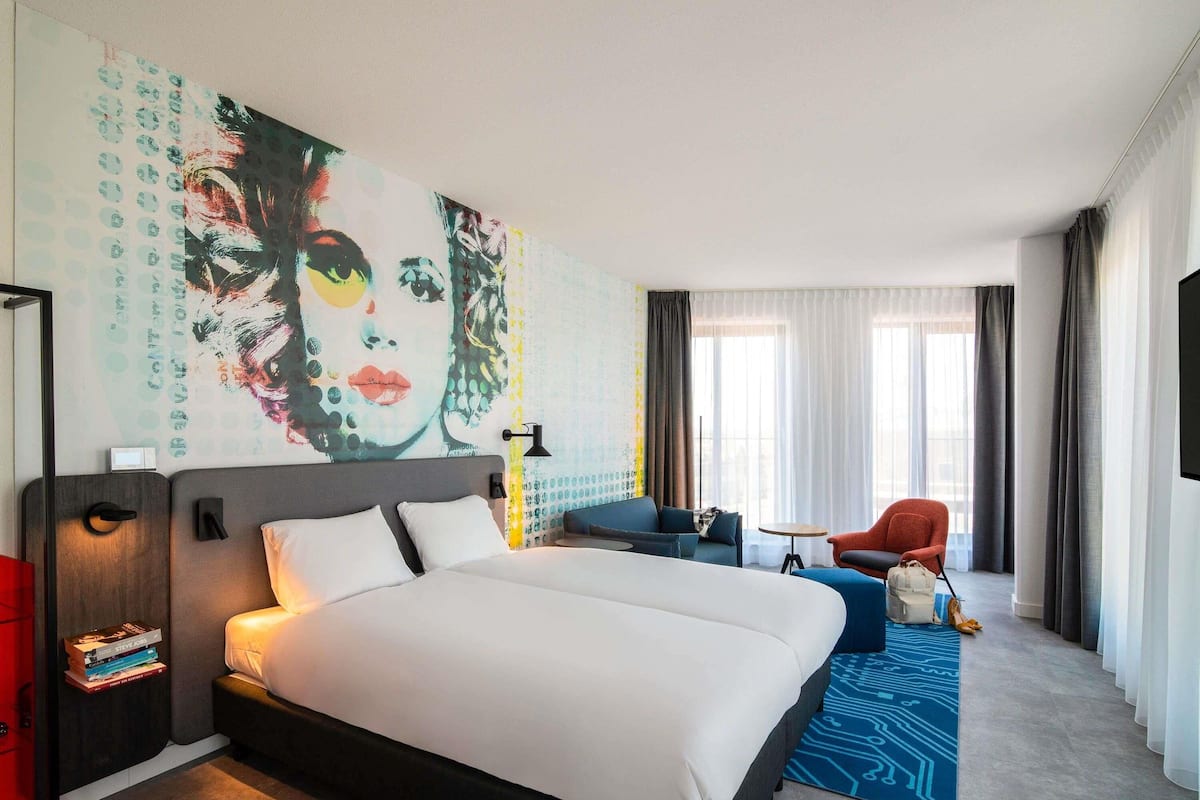Quarto de hotel com uma cama grande, área de estar com sofá e poltrona, televisão e um quadro abstrato de grandes dimensões na parede.