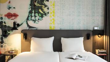 Hotelkamer met een groot bed, twee kussens, een nachtkastje met een lamp, een muur met een kleurrijke muurschildering, en een bijzettafeltje met een telefoon en boeken.