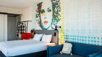 A hotel room with a bed, a sofa, a desk, and a wall with a large abstract painting.