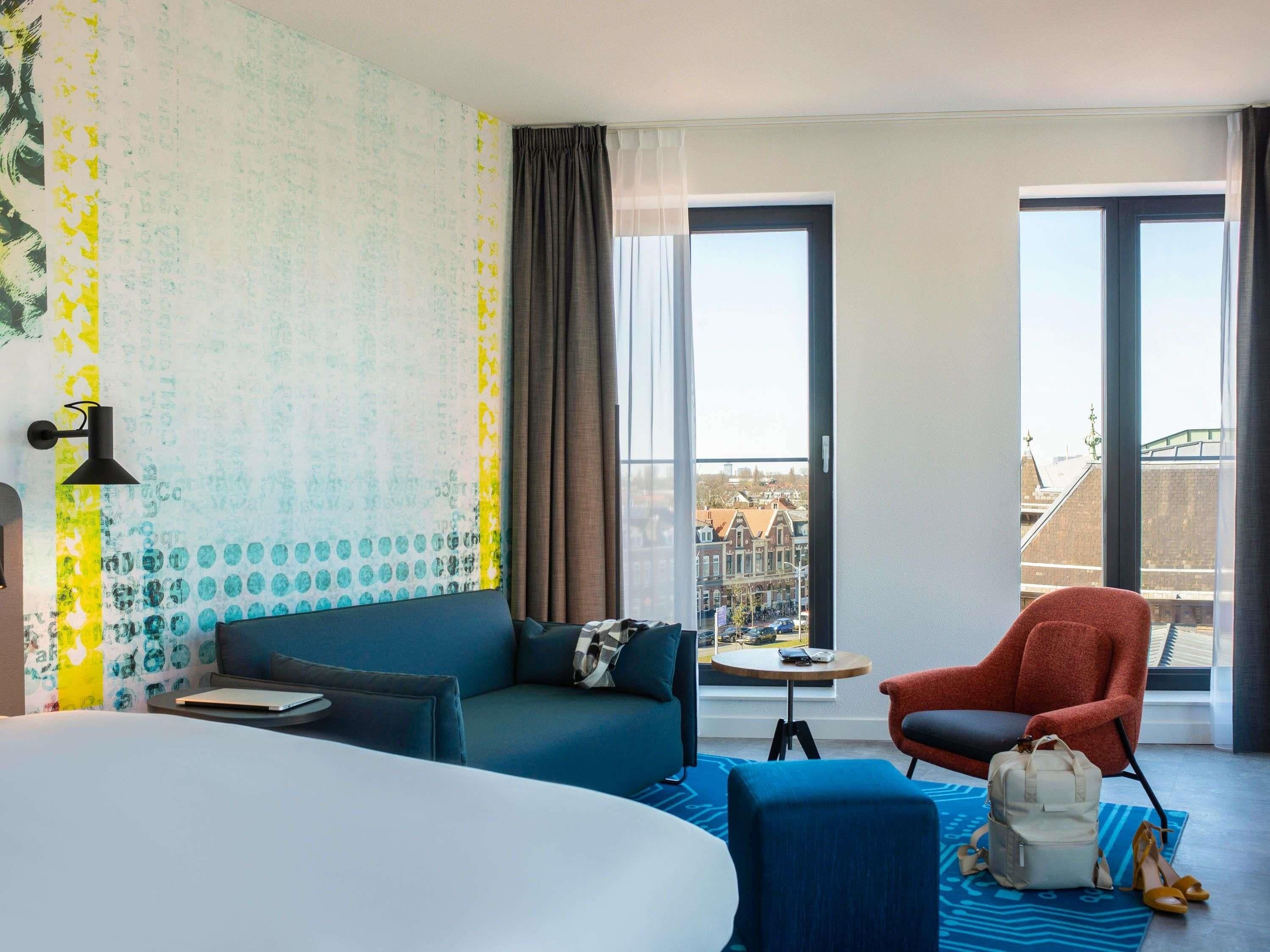 Habitación de hotel con una cama grande, un sofá azul, un sillón rojo, una mesita y vistas a la ciudad a través de amplias ventanas.