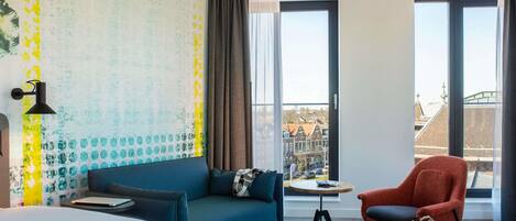 Habitación de hotel con una cama grande, un sofá azul, un sillón rojo, una mesita y vistas a la ciudad a través de amplias ventanas.