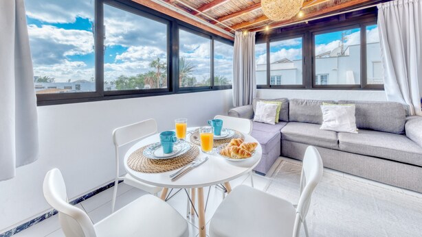 Una habitación moderna con una mesa de comedor dispuesta para el desayuno, un sofá gris y ventanas amplias que dan a vistas del cielo y edificios.