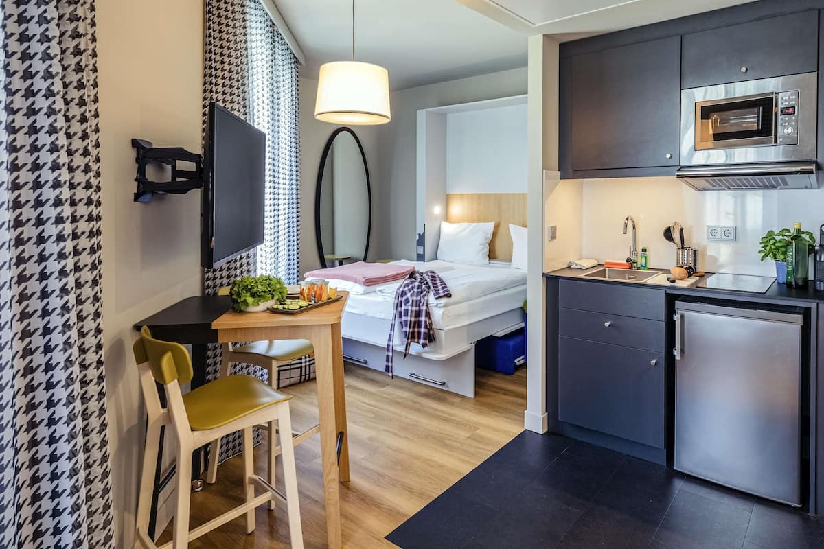 A compact hotel room with a kitchenette, a bed, a dining area, and a TV.