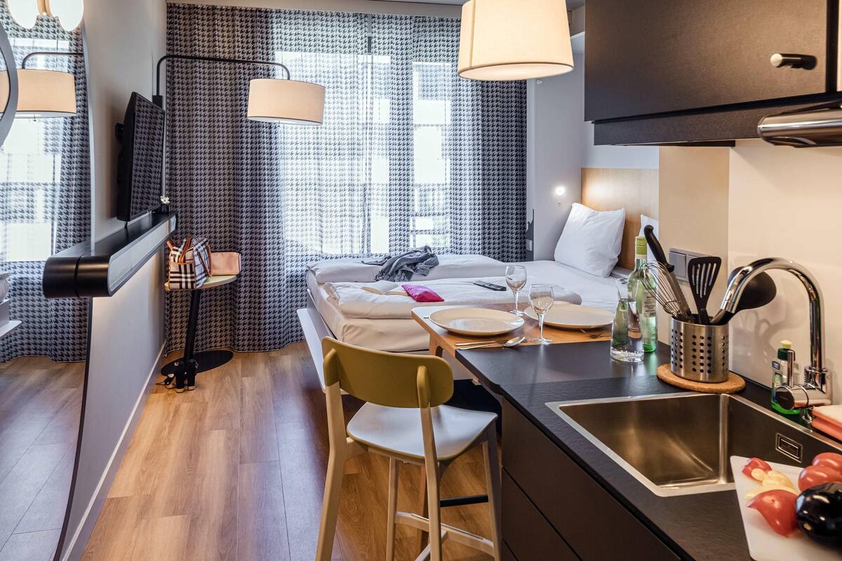A compact hotel room with a bed, bedside table, and a small kitchenette.