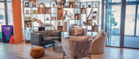 Una sala de estar moderna con un sofá, sillones y una mesa de centro. Hay una estantería llena de diversos objetos y un gran ventanal que proporciona luz natural.
