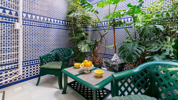 A small, enclosed patio area with a patterned blue and white tiled wall, green plastic chairs, a small table with a bowl of lemons, and a potted plant. - CHIC-ALAMEDA (Sevilla)