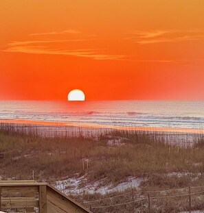 A sunset over a beach with waves, a wooden structure, and sparse vegetation.