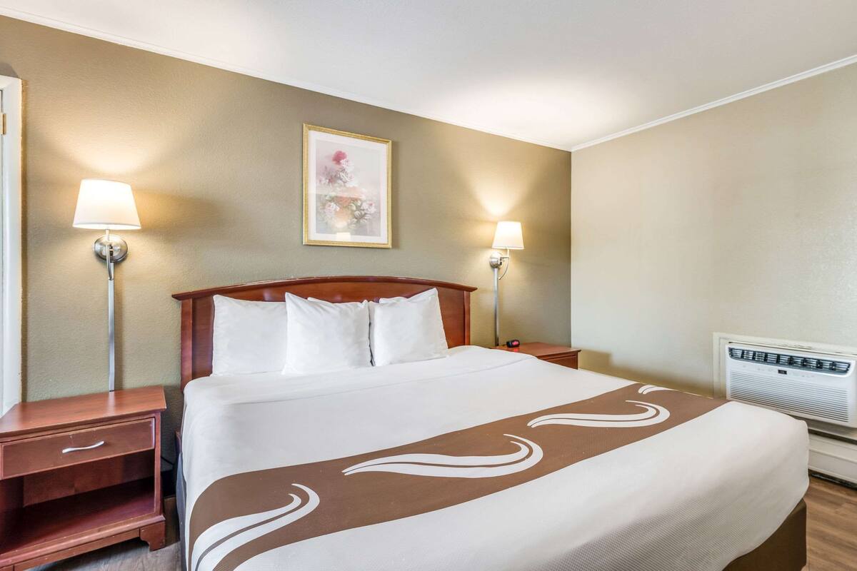 Una habitación de hotel con una cama grande, dos mesitas de noche, aire acondicionado en la pared y dos lámparas de pared.