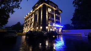 Et hotel i flere etager med oplyst skiltning, en parkeringsplads med flere biler og våd jord der spejler lyset.