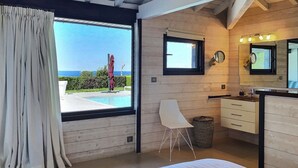 Un dormitorio con un ventanal grande, vistas al mar, techo de madera, una silla blanca y un baño con espejo y lavamanos.
