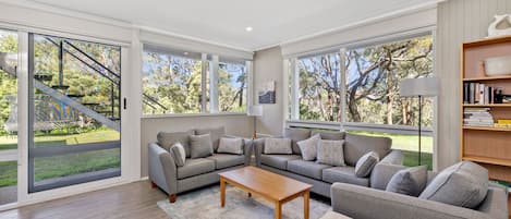 Amplia sala de estar con un sofá seccional gris, una mesa de centro de madera y un ventanal que ofrece vistas de una escalera y vegetación al exterior.