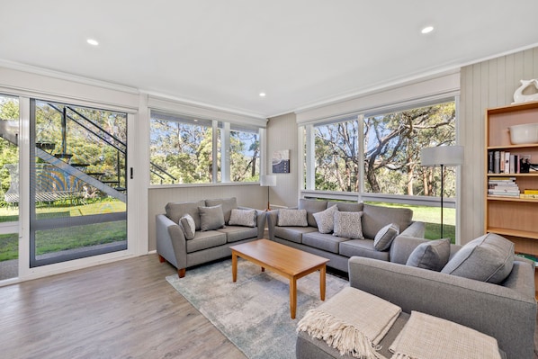 A spacious living room with a grey sectional sofa, a wooden coffee table, and a large window offering a view of a staircase and greenery outside.