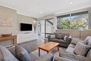 A modern living room with a flat-screen TV, a wooden coffee table, grey sofas, and a large window with a view of trees.
