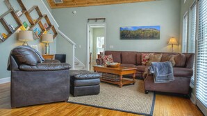 A living room with a brown leather sofa, a wooden coffee table, and a rug. There is a painting on the wall and a staircase leading to another room. - Cozy Cabin with Mountain Views, Pet Friendly, 3 Miles from Downtown Highlands (Highlands)