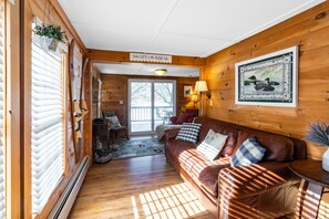 A cozy cabin interior with a wooden floor, a leather sofa, a dining table, and a large window with blinds.