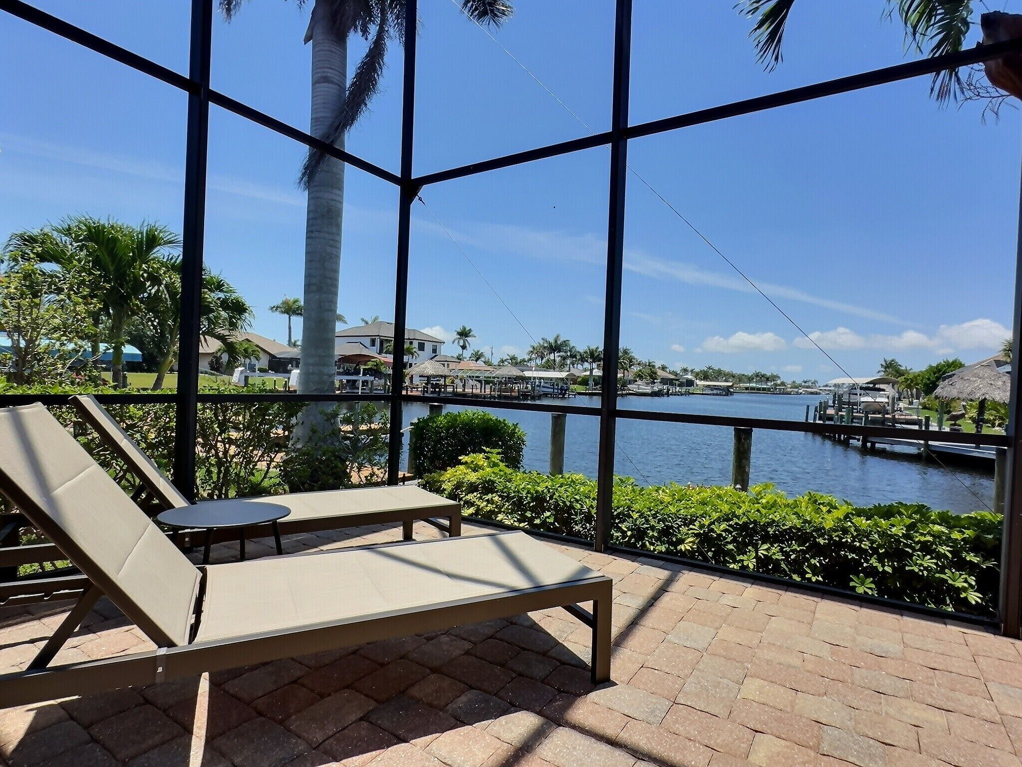 A sunlit outdoor seating area with lounge chairs and a table, overlooking a calm water body with boats and a dock.