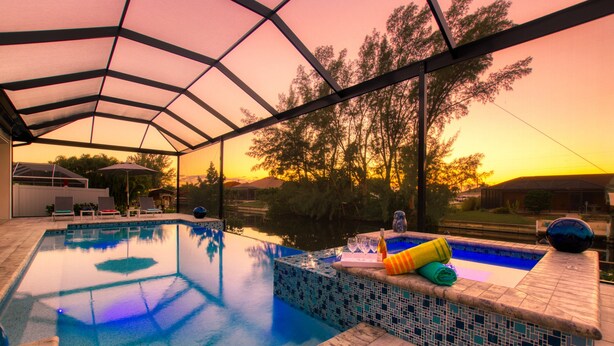An indoor swimming pool with a tiled edge, a covered seating area, and a view of trees and hills. - Modern Waterfront Pool Home with incredible indoor/outdoor spaces (Cape Coral)