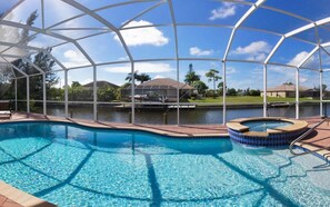 Una piscina con un jacuzzi, rodeada por una zona cubierta con vistas a un lago y casas al fondo.