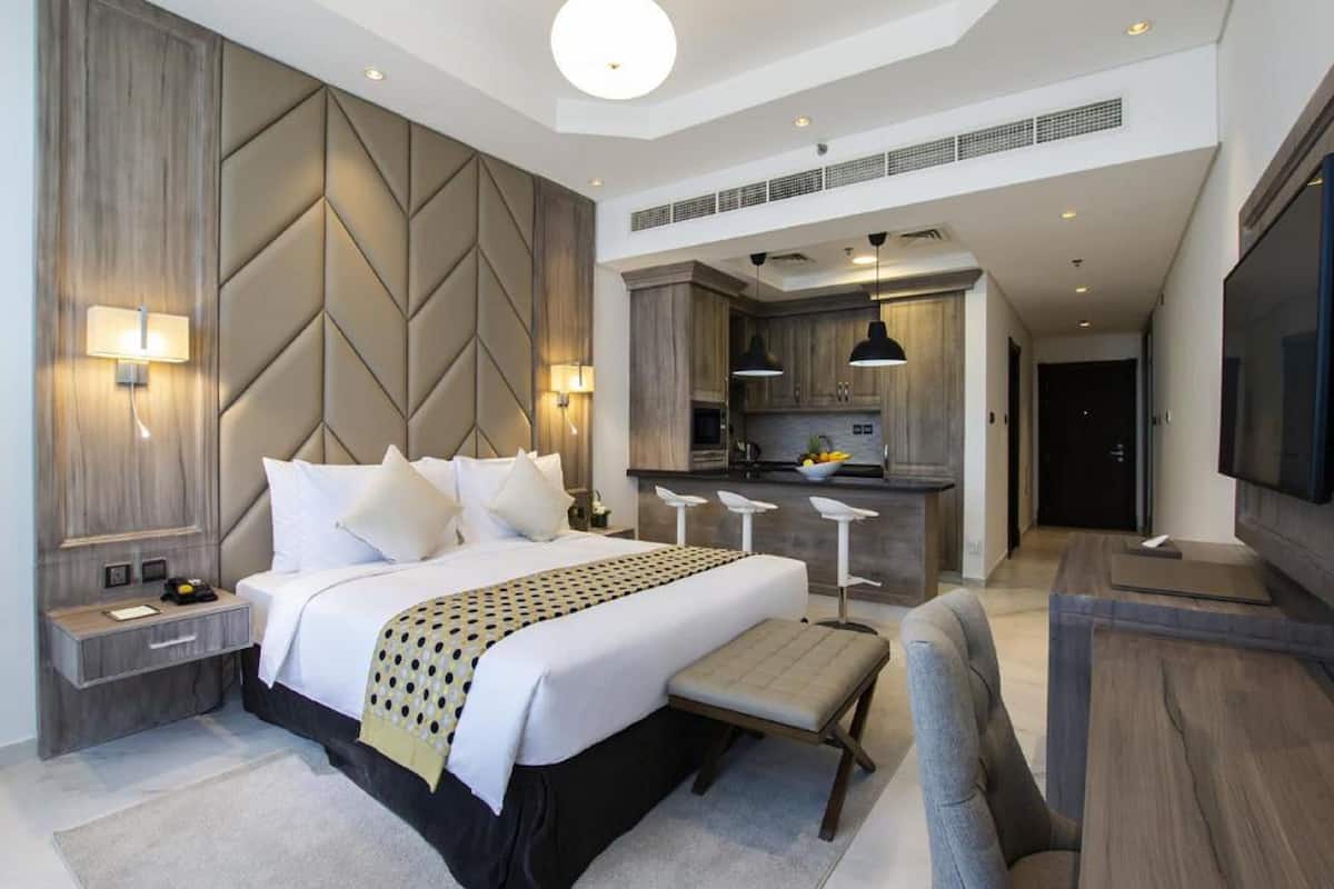 A modern hotel room with a large bed, a desk, a chair, and a kitchenette.