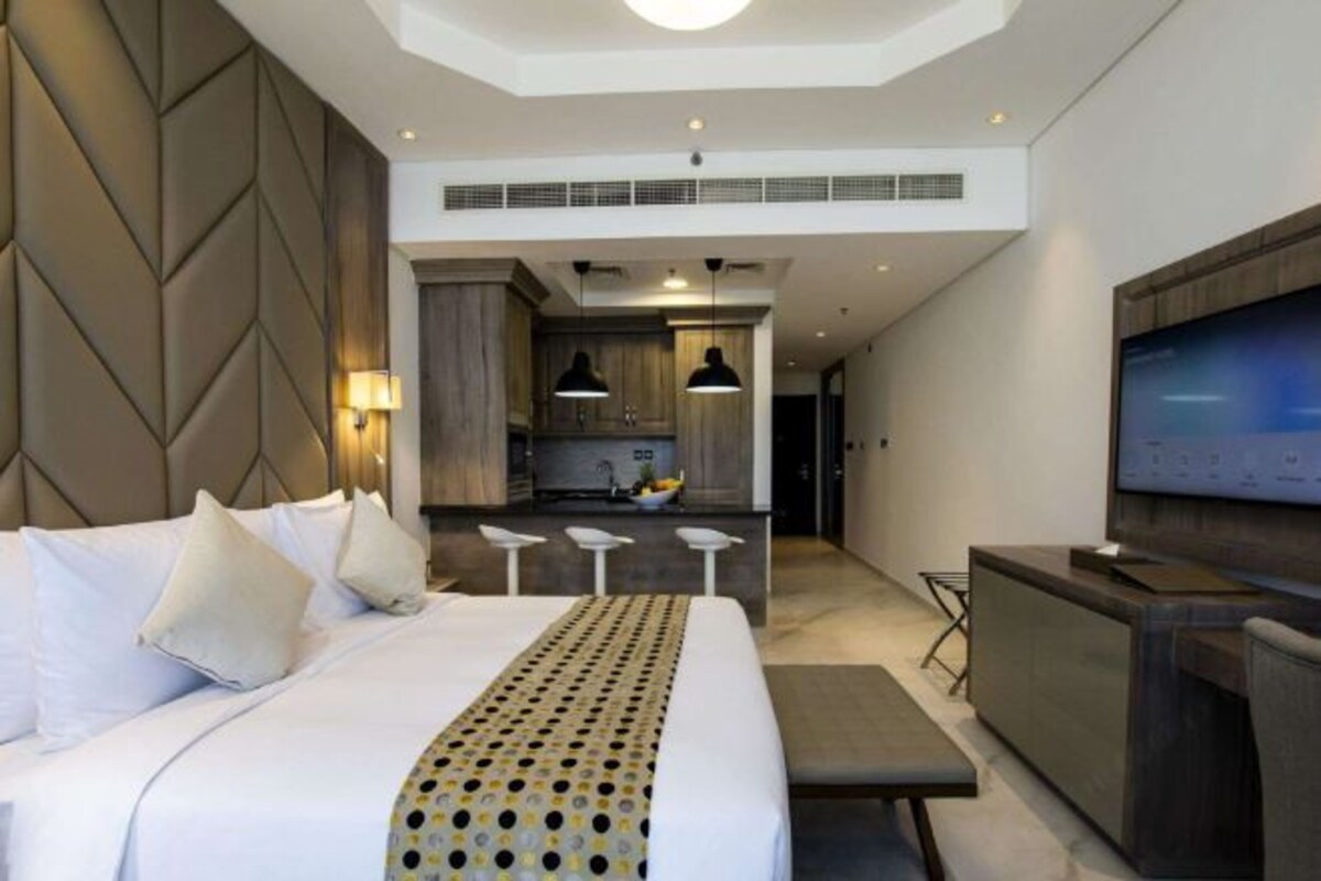 Habitación de hotel moderna con una cama grande, un escritorio, un televisor y una kitchenette al fondo.