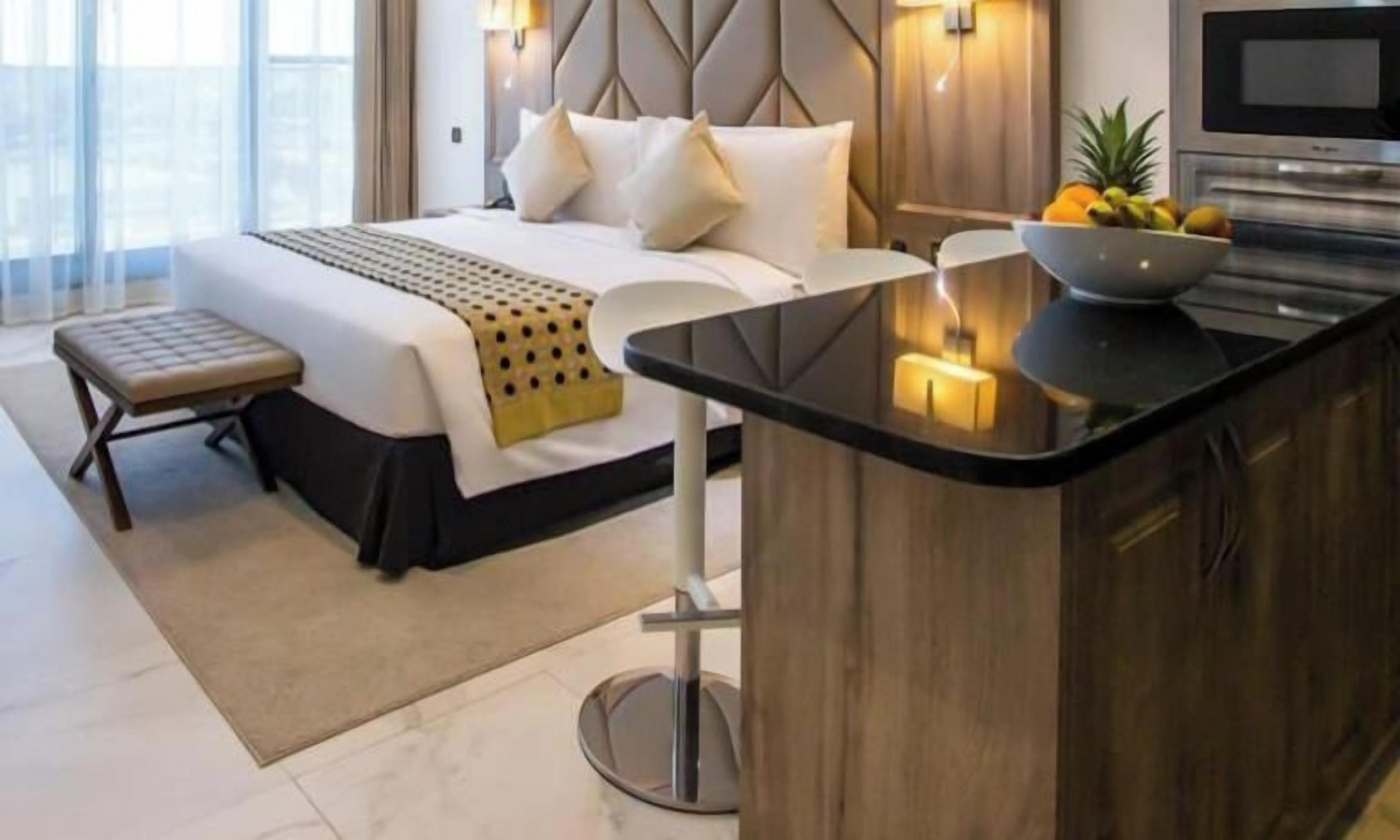 Quarto de hotel moderno com cama, mesa de cabeceira e pequena cozinha equipada com micro-ondas.
