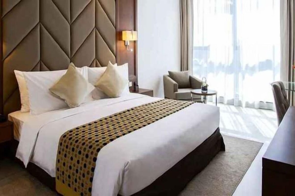 Quarto de hotel com uma cama grande, cabeceira, mesa de cabeceira, uma cadeira e vista para o exterior.