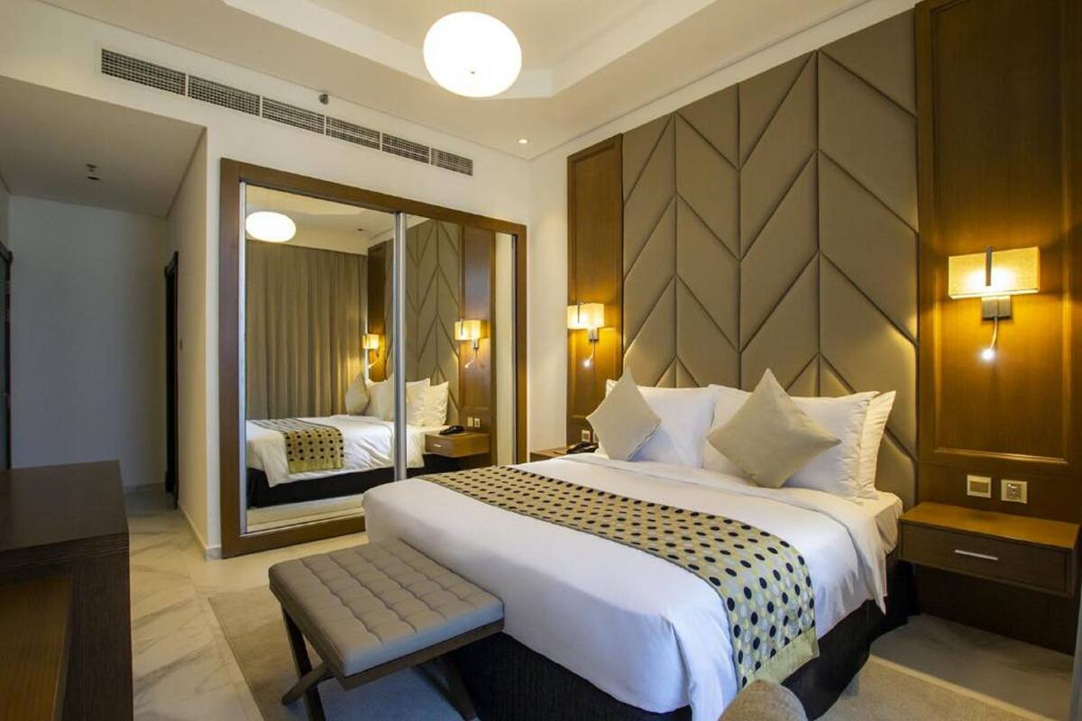 A hotel room with a large bed, a bench, a mirror, and a wall-mounted light fixture.