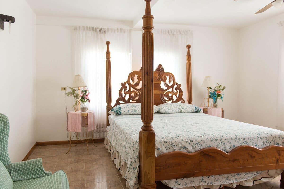 A four-poster bed with a floral bedspread, a green upholstered chair, a small bedside table with a lamp, and a window with sheer curtains.