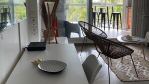 Un espace repas moderne avec une table blanche, des chaises noires et une vue sur les palmiers à travers de grandes fenêtres.