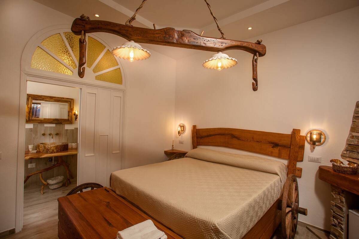 A bedroom with a wooden bed frame, a wooden nightstand with a lamp, a wooden bench, and a white door leading to a bathroom.