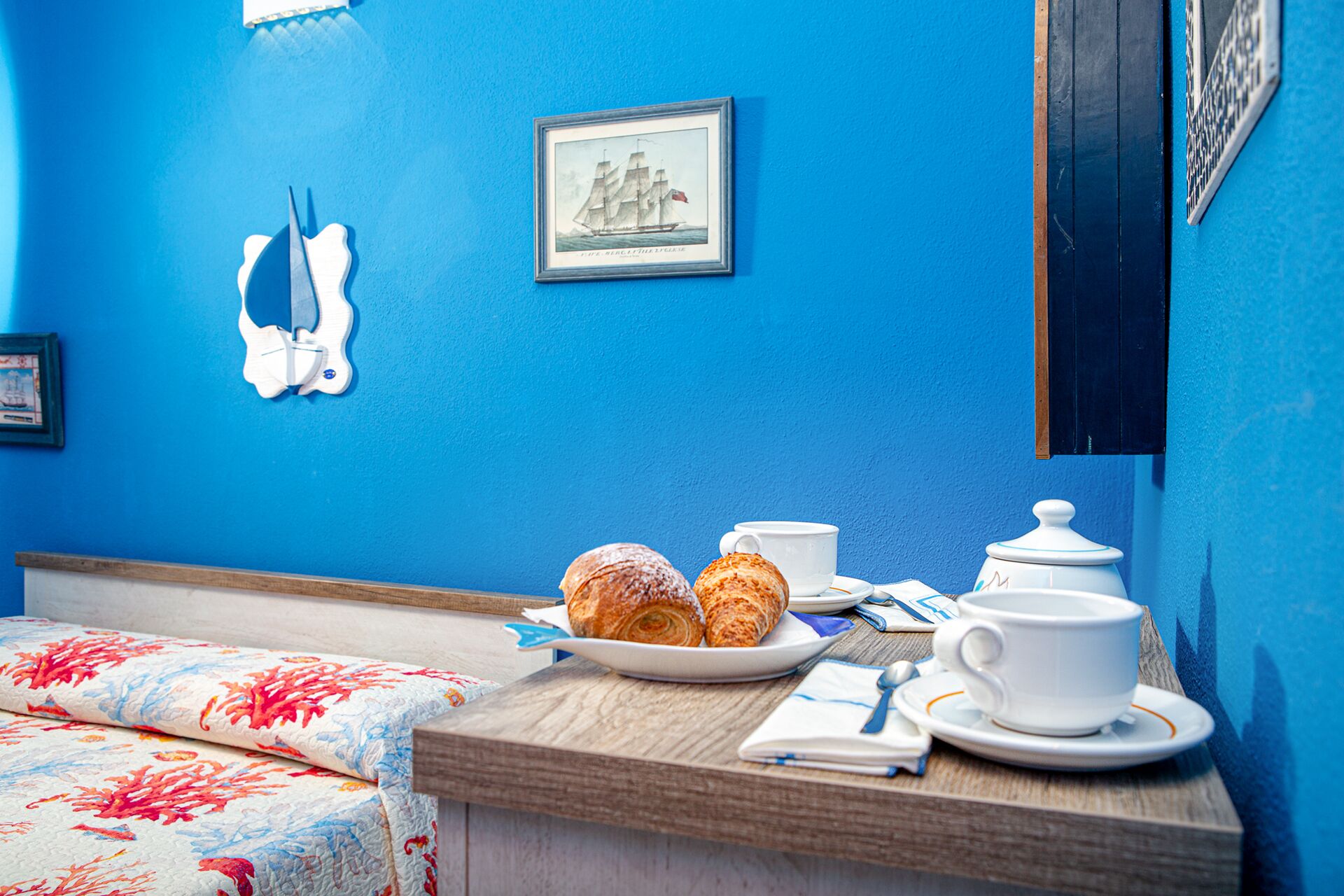 Una habitación con pared azul, cama con colcha estampada, mesita de noche de madera con un plato de croissants, una taza blanca, una tetera blanca y un cuadro con una imagen de un barco.