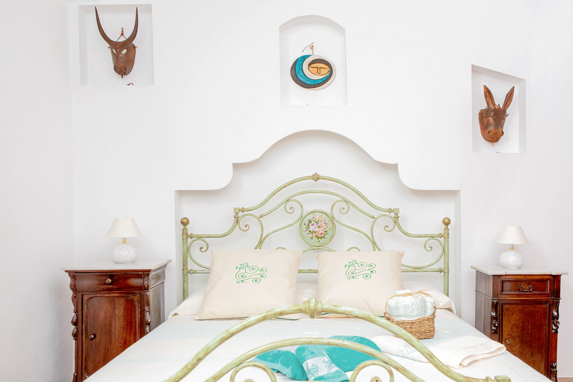 A bed with a green metal frame, two pillows with green embroidery, and a bedside table with a lamp.