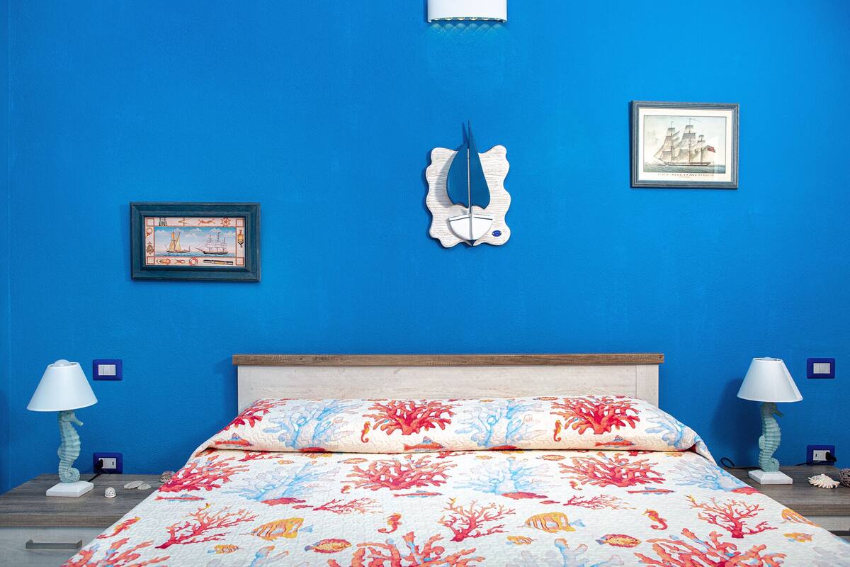 A bedroom with a blue wall, a bed with a coral-patterned bedspread, two nightstands with lamps, a framed picture, and a decorative wall hanging.