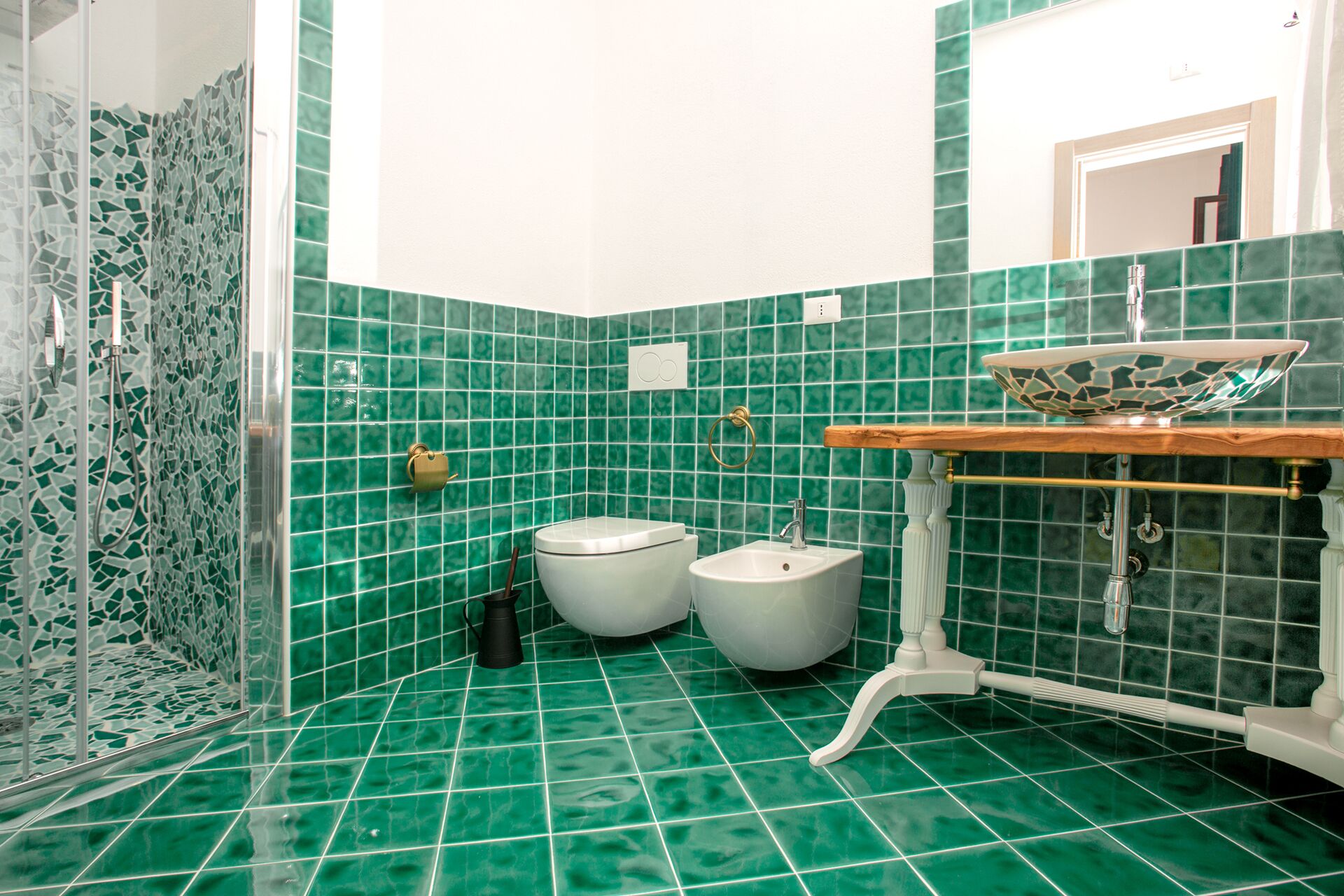 Un baño con mosaicos verdes, un inodoro blanco, un bidé y un lavamanos con encimera de madera.
